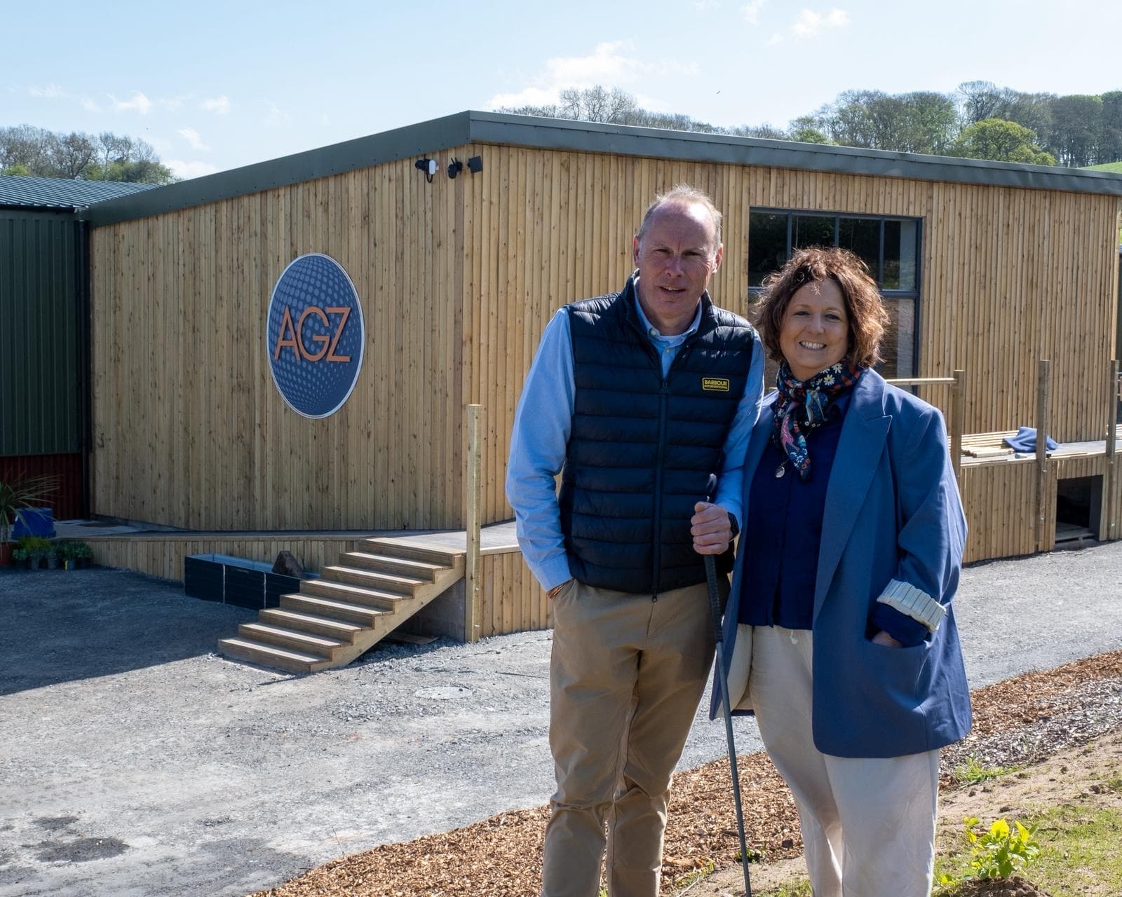 Robert and Donna Ashbrook standing outside the Ashbrook Golf Zone clubhouse on Bradeley Green Lane, Whitchurch — the AGZ logo on the timber-clad building