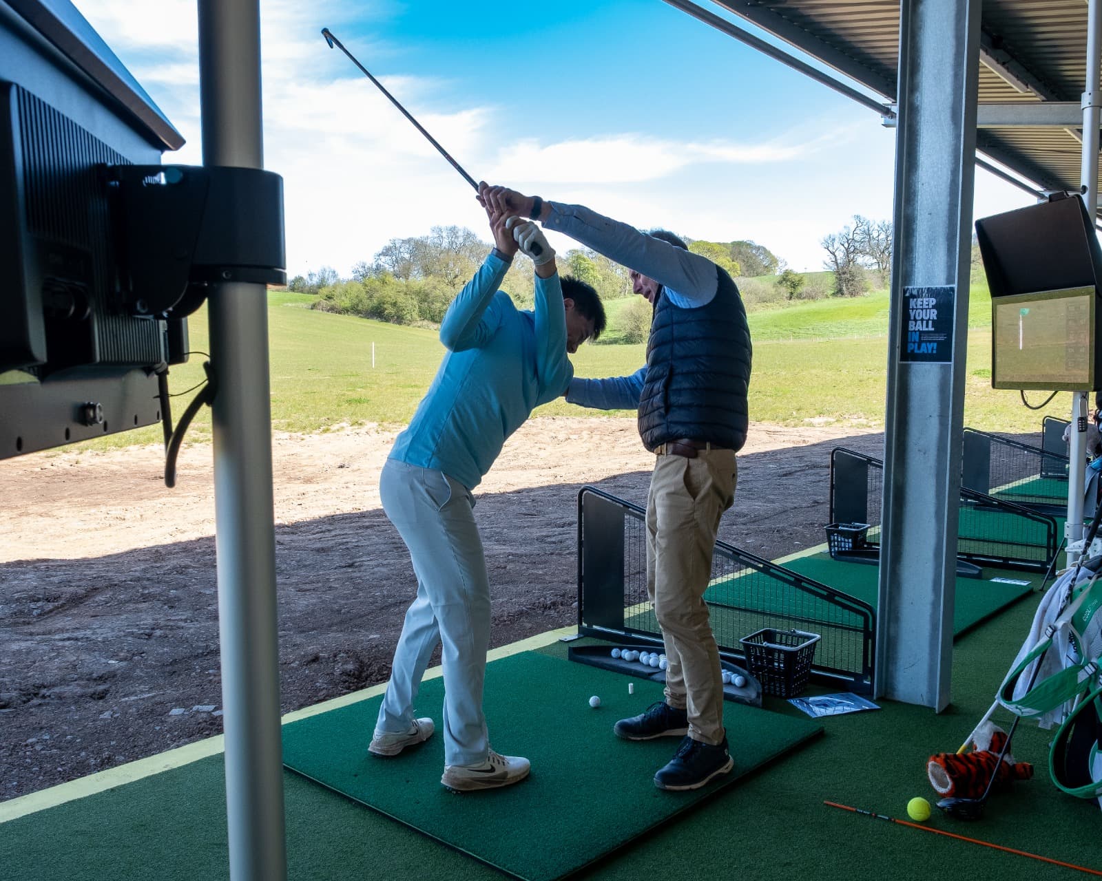 A coach guiding a student through their swing at an Ashbrook Golf Zone Toptracer bay, the practice field beyond